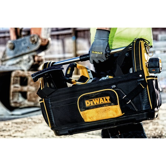 Close-up of a DEWALT-branded tool bag filled with various tools, being carried by a person wearing work gloves and a safety vest at a construction site.