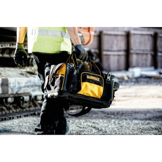 A person wearing work gloves and a reflective vest is holding a DEWALT-branded yellow and black tool bag at a construction site.