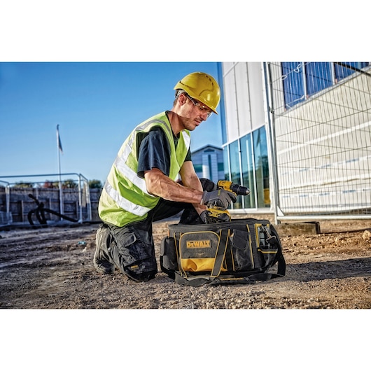 A construction worker wearing a yellow safety helmet and reflective vest is kneeling on the ground outside a building site, using a DEWALT cordless drill next to a black and yellow DEWALT tool bag.