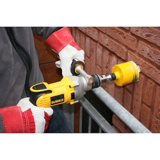 A person wearing protective gloves is using a yellow DEWALT power drill with a hole saw attachment to drill into a brick wall.