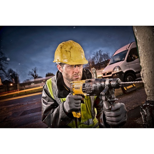 A construction worker wearing a yellow hard hat and reflective jacket uses a DEWALT hammer drill outdoors, with a white van and blurred lights in the background.