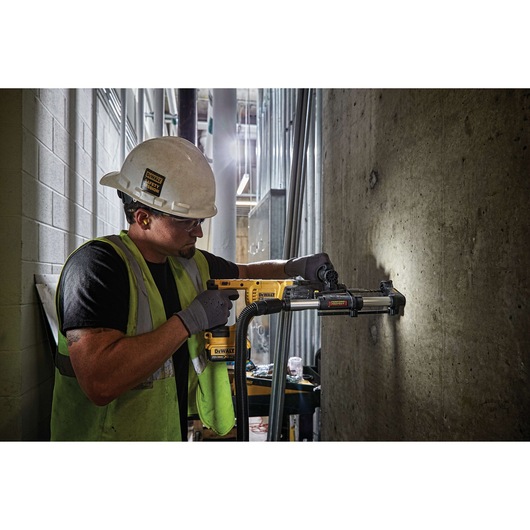 A construction worker wearing a hard hat and safety vest operates a yellow DEWALT power tool with a dust extraction attachment, drilling into a concrete wall in an industrial setting.