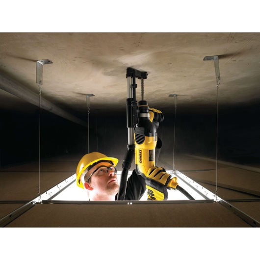 A person wearing safety gear and gloves uses a yellow DEWALT power tool to drill into a ceiling inside a construction site, viewed from an opening in the ceiling tiles.