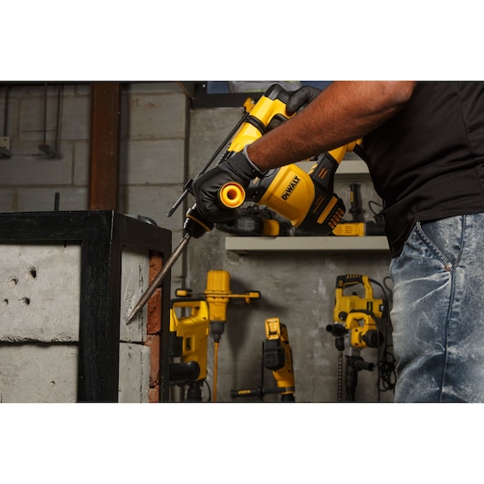 A person wearing gloves uses a DEWALT rotary hammer drill to chisel concrete blocks in a workshop. Other yellow DEWALT power tools are visible in the background.