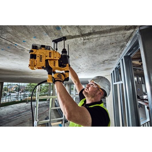 A person wearing a safety helmet and vest is using a DEWALT power tool to drill into a concrete ceiling at a construction site. The DEWALT tool is held overhead and appears to be a rotary hammer drill.