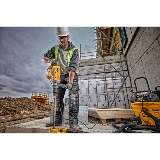 A construction worker wearing a safety helmet and high-visibility vest operates a DEWALT rotary hammer drill (SKU: D25733K) on a building site. The worker's face is blurred, and there is construction equipment, scaffolding, and insulation materials visible in the background.