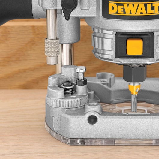 Close-up image of a DEWALT plunge router being used on a piece of wood. The router features metallic parts, adjustment knobs, and a visible cutting bit actively engaged with the surface.