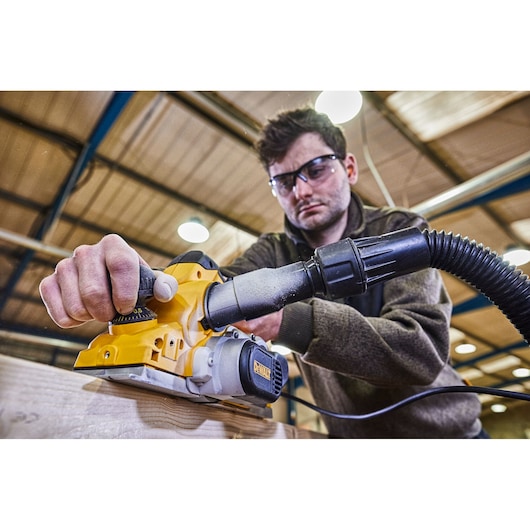 A person using a DEWALT D26500 electric planer on a piece of wood in an indoor workshop. The person's face is blurred, and the planer is connected to a hose for dust extraction.