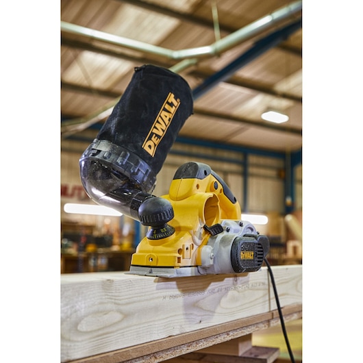 A DEWALT electric hand planer is shown in use, smoothing a wooden board in a workshop. The planer has a dust bag attached to the top to collect wood shavings.