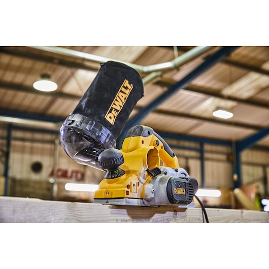 A yellow DEWALT electric planer with a dust bag attached, placed on a wooden surface inside a workshop.