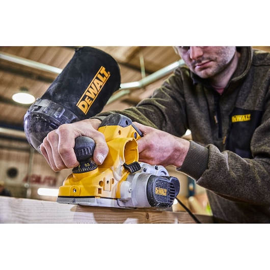 A person using a DEWALT D26500 electric planer on a wooden surface in a workshop setting. The person’s face is obscured, and the DEWALT logo is visible on the dust bag and their clothing.