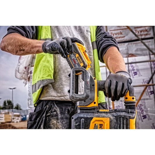 Close-up of a person wearing gloves and a high-visibility vest, inserting a DEWALT DCB548 XR FLEXVOLT battery into a compatible DEWALT power tool at a construction site.