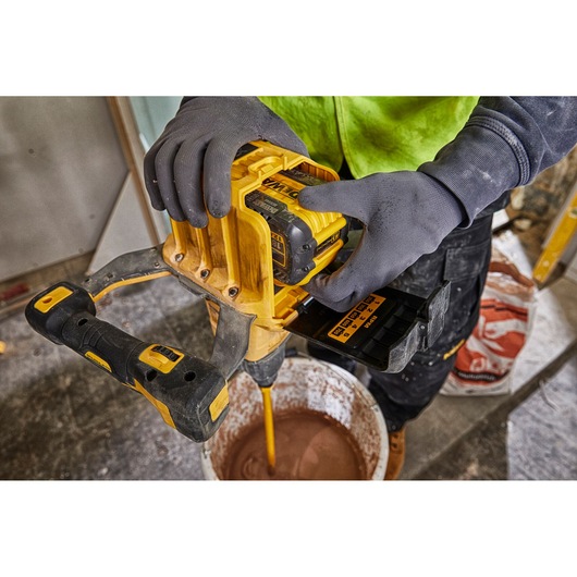 A person wearing gloves uses a yellow DEWALT cordless mixer with a DEWALT DCB548 battery to mix material in a bucket on a construction site.