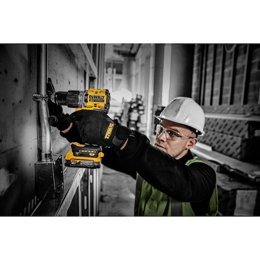 A person wearing safety gear, including gloves and a white hard hat, uses a yellow DEWALT cordless drill with a POWERSTACK