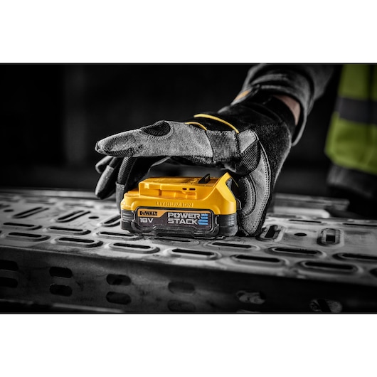 Gloved hand holding a DEWALT 18V POWERSTACK battery on a metal surface at a worksite.