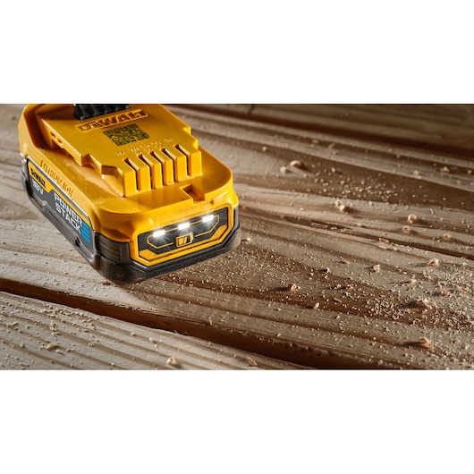 DEWALT DCBP034 battery pack on a wooden surface with sawdust, LED lights visible on the front.