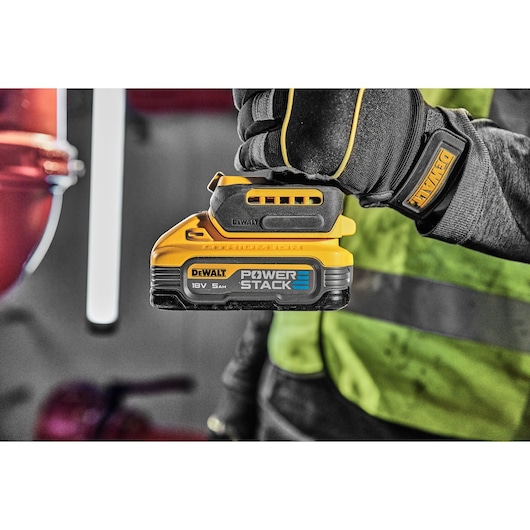 A gloved hand holds a DEWALT POWERSTACK 18V 5.0Ah battery pack in a workshop setting.