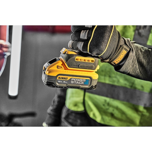 Person wearing gloves holds a DEWALT POWERSTACK 18V 5Ah lithium-ion battery in a work environment.