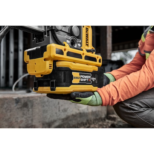 A person wearing work gloves and an orange jacket is holding a DEWALT Power Shift 554 tool or battery unit on a construction site.