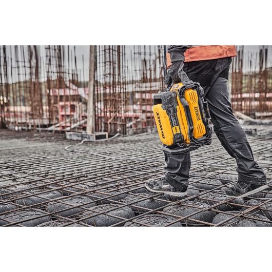 Person wearing work gloves and construction clothing carrying a yellow DEWALT power tool battery on a construction site with metal rebar and unfinished structures.