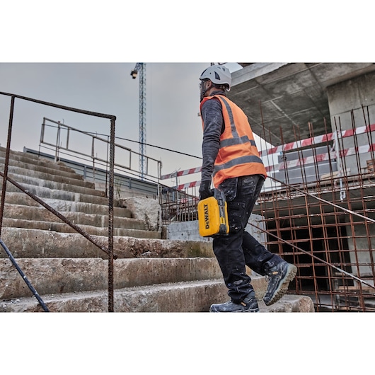 A person wearing safety gear and an orange reflective vest walks up concrete stairs at a construction site, holding a yellow DEWALT tool in their hand.