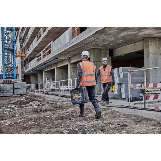 Two construction workers wearing orange safety vests and helmets are walking on a construction site. One is carrying a black DEWALT toolbox, and the other is holding a yellow tool. Concrete blocks and scaffolding are visible in the background.