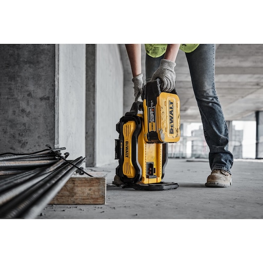 A person wearing gloves and work boots is using a yellow DEWALT portable power station on a construction site. Metal rods and concrete surfaces are visible in the background.