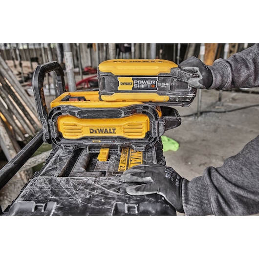 A person wearing gloves installs a yellow and black DEWALT Power Shift battery pack onto a DEWALT tool at a construction site. The battery and tool surfaces are dusty, and construction materials are visible in the background.