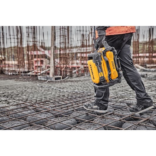 A person wearing construction gear is carrying a yellow DEWALT portable tool on a construction site with steel rebar and unfinished building structures in the background.