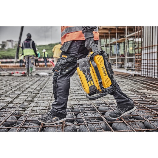 A person in workwear and gloves carrying a yellow and black DEWALT battery charger while walking on a construction site with rebar and concrete spheres. Other workers and building scaffolding are visible in the background.