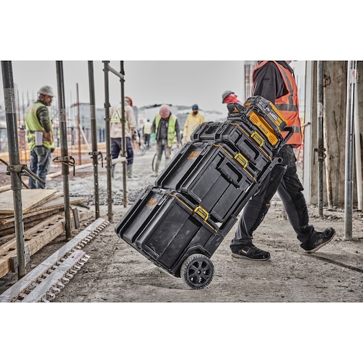 A worker pushes a DEWALT rolling stack of black and yellow tool storage boxes on a construction site, surrounded by scaffolding and other workers.
