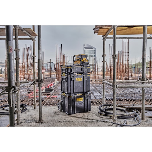 A stack of DEWALT black tool storage boxes on wheels is placed on a construction site, surrounded by metal scaffolding and rebar. The boxes feature yellow DEWALT branding and are suitable for storing and transporting tools.