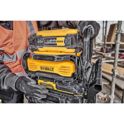 Close-up of a DEWALT Power Shift 54V battery attached to a DEWALT ToughSystem storage unit, held by a person wearing safety gloves and an orange high-visibility vest at a construction site.