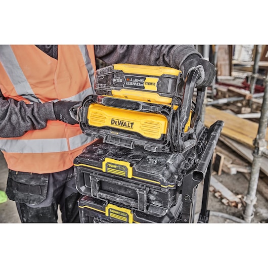 A person wearing an orange safety vest and black gloves is handling stacked DEWALT ToughSystem toolboxes with a yellow DEWALT Power Shift battery and charger on top at a construction site.