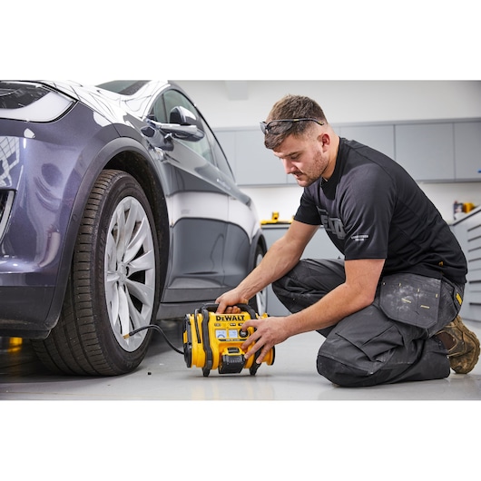 A person kneeling on the floor is using a DEWALT DCC018N portable air compressor to inflate the tire of a parked car inside a garage or workshop. The person's face is blurred for privacy.