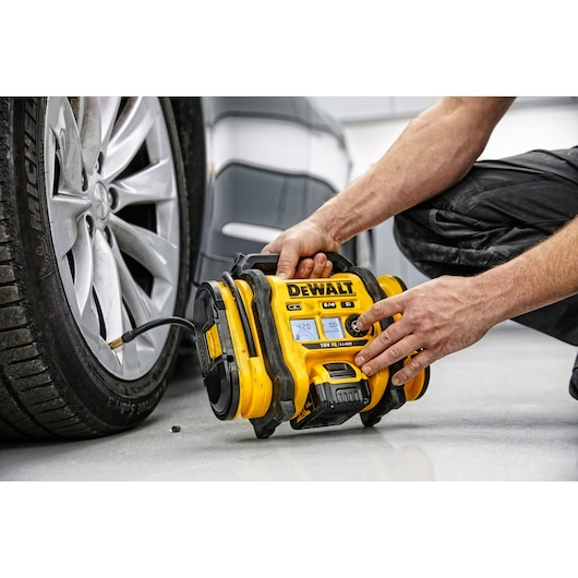 A person using a yellow DEWALT portable air compressor to inflate the tire of a car. The device is connected to the tire valve and placed on the ground next to the vehicle.