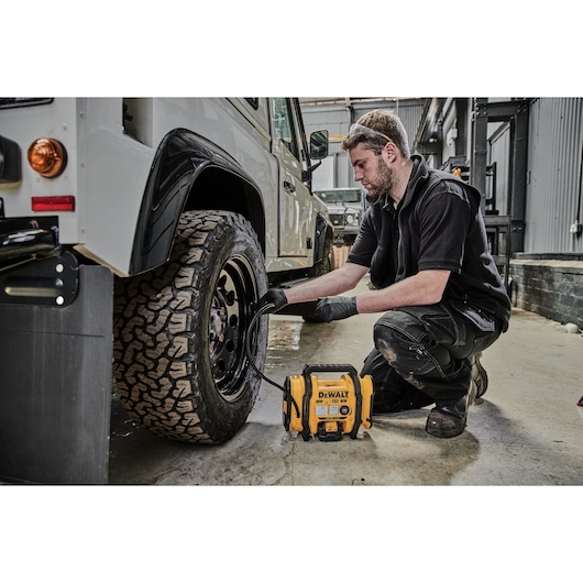 A person kneels next to a rugged vehicle tire in an industrial workshop, using a DEWALT DCC018N portable air compressor to inflate the tire.