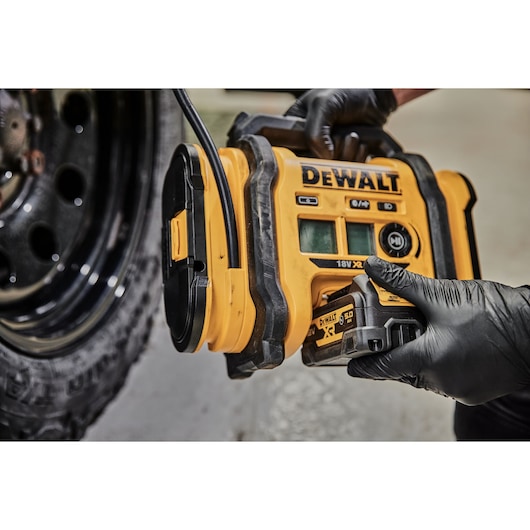 A close-up view of a person wearing black gloves using a DEWALT portable digital air compressor (model DCC018N) to inflate a car tire.