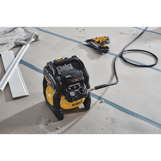 DEWALT cordless air compressor with hose on a construction floor, next to a nail gun and white trim pieces.
