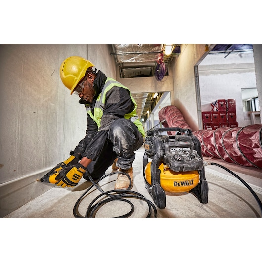 A construction worker wearing a yellow hard hat and safety vest is kneeling on a building site, using a power tool attached to a DEWALT cordless air compressor model DCC1054N. The compressor is placed on the floor, and the worker is working along a wall. The surrounding area shows industrial equipment and materials.