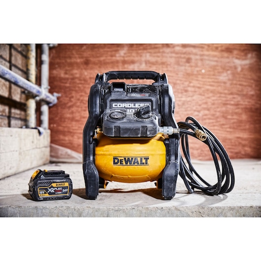 A DEWALT cordless air compressor with a yellow tank and black casing, shown with a detachable battery pack. Both items are placed on a concrete surface in a workshop setting.