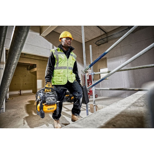 A construction worker wearing a yellow hard hat, high-visibility vest, gloves, and boots is carrying a DEWALT portable air compressor inside a building under construction. The worker's face is blurred for privacy.