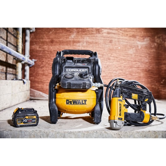 A DEWALT cordless air compressor with a yellow tank and black handle is placed on a concrete surface. Next to it is a DEWALT battery and an air tool with attached hose, all in a workshop setting.
