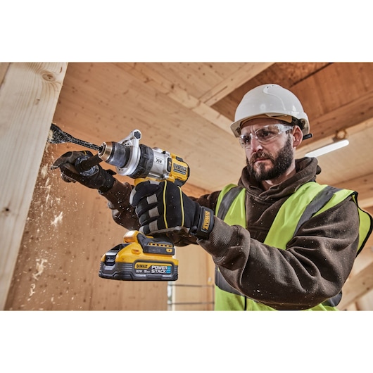 A person wearing safety gear uses a DEWALT cordless drill (model DCD1007H2T) to drill into a wooden beam, with wood shavings