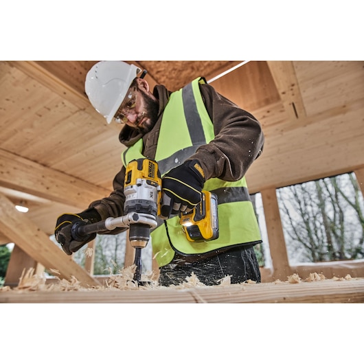 A construction worker wearing a safety helmet and high-visibility vest is using a DEWALT cordless drill to bore holes into a