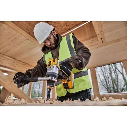 A person wearing a white safety helmet and high-visibility vest is using a DEWALT cordless drill to bore a hole in a wooden
