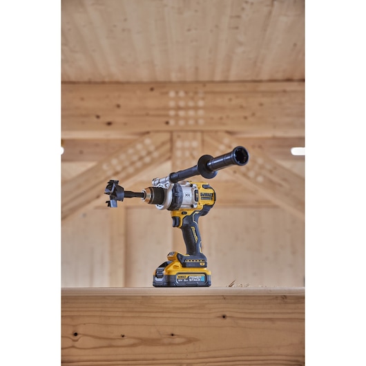 Yellow and black cordless drill with a side handle and bit, placed on a wooden beam indoors.