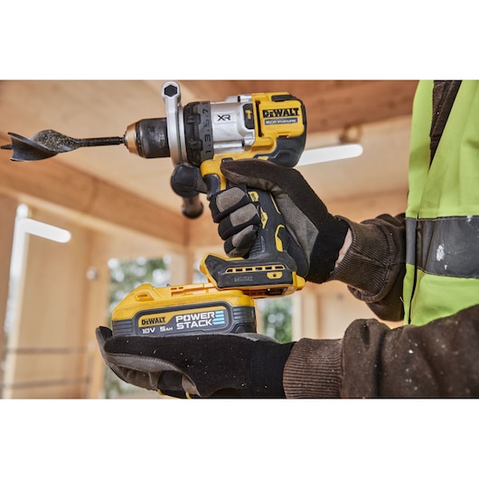A DEWALT cordless drill, model DCD1007NT, is being held by a person wearing work gloves and a high-visibility jacket. The drill features a large auger bit and a Power Stack 18V 5Ah battery. Suitable for professional woodworking or construction tasks.
