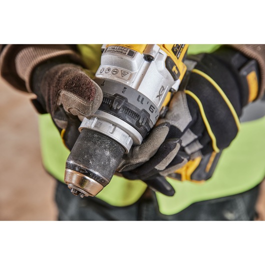 Close-up of a person wearing work gloves and a high-visibility vest, holding a DEWALT cordless drill, focusing on the chuck and adjustment ring.