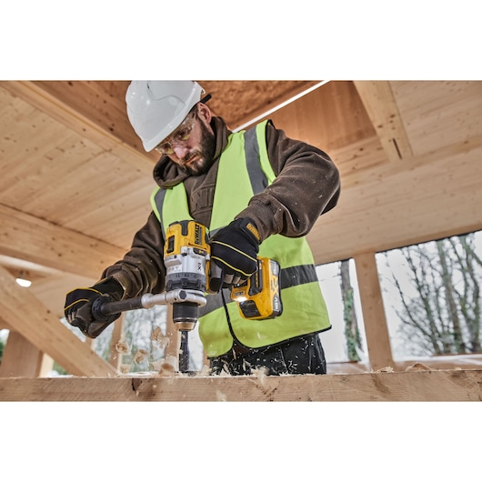 A person wearing a safety helmet and high-visibility vest is using a DEWALT cordless drill to bore a hole into a wooden beam on a construction site.